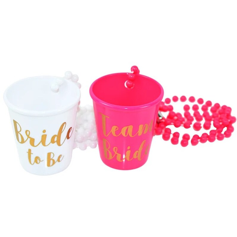 Pu&ograve; includere: Due bicchieri da shot in plastica con scritta dorata. Uno &egrave; bianco con il testo "Bride to Be" e l'altro &egrave; rosa con il testo "Team Bride". &Egrave; incluso un collier di perline rosa.