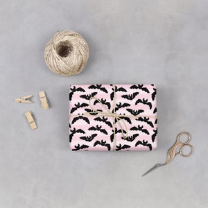 Op de afbeelding: Een roze en zwart cadeaupapier met een patroon van vleermuizen. Het cadeau is ingepakt met touw en vastgebonden met een strik.
