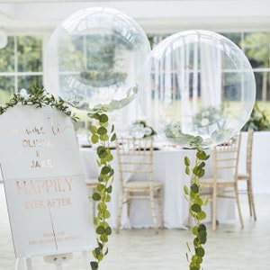 Puede incluir: Dos globos transparentes con vegetación en su interior, decorados con una guirnalda verde, y un cartel blanco con el texto "Bienvenido a Olivia & Jake Happily Ever After Gracias por celebrar con nosotros".