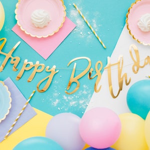 May include: A blue and white background with a gold foil "Happy Birthday" banner. There are yellow, blue, and pink balloons, as well as pink and blue paper plates with white marshmallows on them.