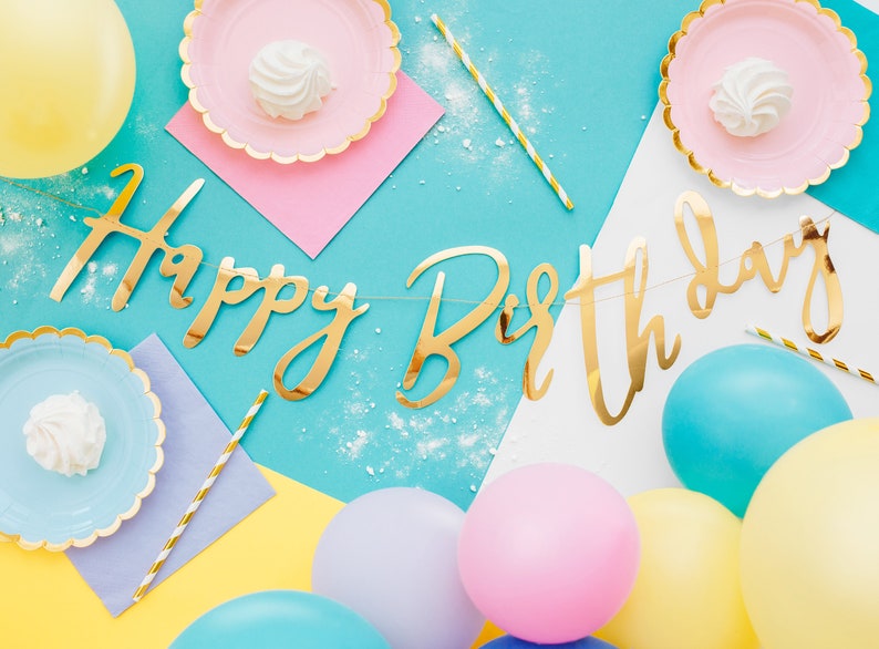 May include: A blue and white background with a gold foil "Happy Birthday" banner. There are yellow, blue, and pink balloons, as well as pink and blue paper plates with white marshmallows on them.