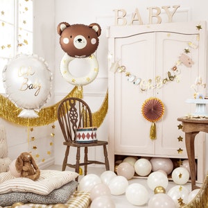 May include: A baby shower party decoration set with a brown bear balloon, a white balloon with the text "Oh Baby", gold fringe, and a banner that says "Oh Baby". There are also gold stars, a wooden chair, and a table with a cake and other decorations.