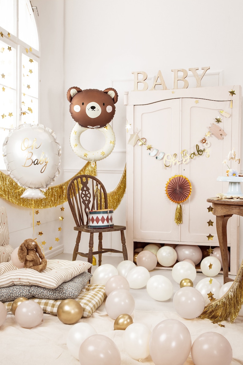 May include: A baby shower party decoration set with a brown bear balloon, a white balloon with the text "Oh Baby", gold fringe, and a banner that says "Oh Baby". There are also gold stars, a wooden chair, and a table with a cake and other decorations.