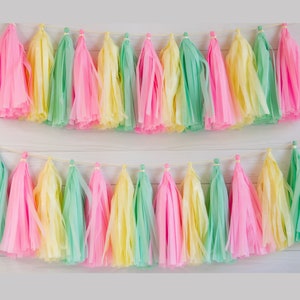 Pode incluir: Duas fileiras de borlas de papel coloridas em tons pastel de rosa, amarelo e verde s&atilde;o enfiadas em um cord&atilde;o fino e de cor clara. As borlas s&atilde;o dispostas sobre um fundo de madeira branca, criando uma faixa festiva e decorativa.