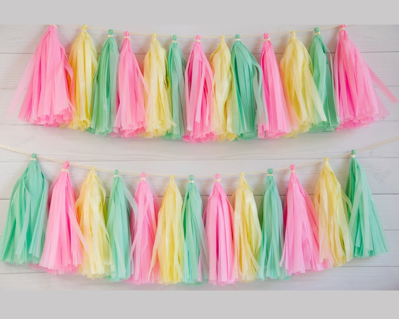 Pode incluir: Duas fileiras de borlas de papel coloridas em tons pastel de rosa, amarelo e verde s&atilde;o enfiadas em um cord&atilde;o fino e de cor clara. As borlas s&atilde;o dispostas sobre um fundo de madeira branca, criando uma faixa festiva e decorativa.
