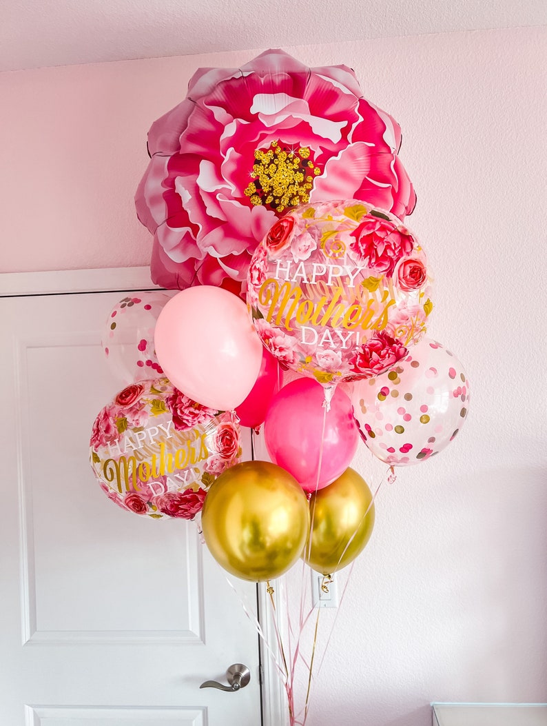 May include: A bouquet of pink, gold, and clear balloons with floral and confetti patterns. The balloons spell out "Happy Mother's Day!" and are tied together with pink ribbons. A large pink flower balloon is at the top of the bouquet.