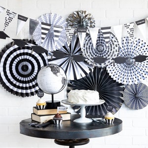 Puede incluir: Decoraciones para fiesta de graduación en blanco y negro con un abanico de papel a rayas blanco y negro, un globo terráqueo y un pastel blanco sobre un soporte para pastel blanco. Las decoraciones incluyen una pancarta con la palabra "Felicidades" y abanicos de papel blanco y negro.