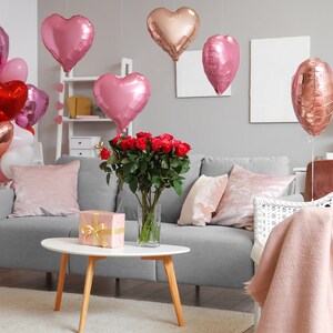 May include: A living room decorated for Valentine's Day with a gray sofa, a white wicker chair, a bouquet of red roses, and a variety of pink and red heart-shaped balloons.