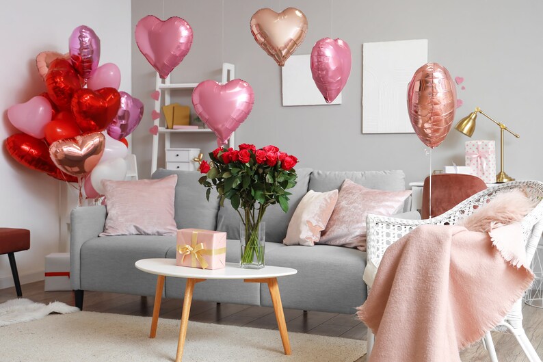 May include: A living room decorated for Valentine's Day with a gray sofa, a white wicker chair, a bouquet of red roses, and a variety of pink and red heart-shaped balloons.