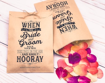 May include: Two brown paper bags with black text that reads "When the Bride and Groom Walk Your Way, Toss This in the Air and Shout Hooray" and "When the Bride and Groom Walk Your Way, Toss This in the Air and Shout Hooray". One bag is open and spilling out pink, white, and purple rose petals.