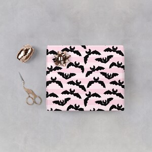 Op de afbeelding: Roze en zwart inpakpapier met een herhalend patroon van vleermuizen. De vleermuizen zijn zwart en vliegen in horizontale positie. De achtergrond is roze met witte strepen.