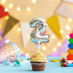 May include: A silver number two balloon on top of a frosted cupcake with sprinkles. The cupcake is on a blue table with party decorations in the background.