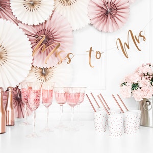 May include: A bridal shower party setup with a gold "Miss to Mrs" banner, pink and white paper fans, rose gold champagne flutes, mini rose gold bottles, pink carnations in a silver bucket, and white cups with pink polka dots.