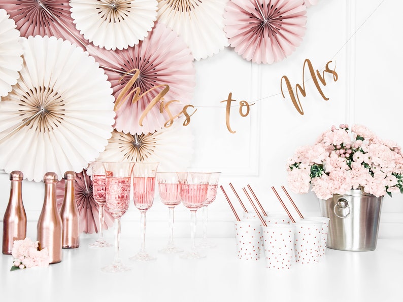 May include: A bridal shower party setup with a gold "Miss to Mrs" banner, pink and white paper fans, rose gold champagne flutes, mini rose gold bottles, pink carnations in a silver bucket, and white cups with pink polka dots.
