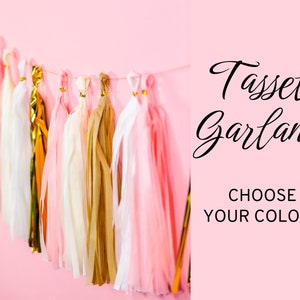 Pode incluir: Uma guirlanda decorativa de borlas em tons de rosa, dourado e branco pendurada em um fundo rosa. A guirlanda apresenta v&aacute;rias borlas com detalhes dourados. O texto &agrave; direita diz "Tassel Garland, Choose Your Colors."