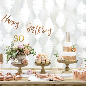 May include: A rose gold 30th birthday party table setting with a white cake decorated with rose gold accents, macarons, and rose gold party supplies.