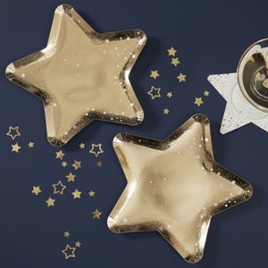 May include: Two gold star-shaped paper plates with a glittery finish. The plates are on a dark blue background with gold glitter confetti.