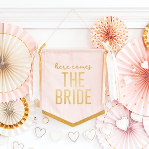 K&ouml;nnte beinhalten: Rosa und goldene Papierf&auml;cher mit goldenen Verzierungen und wei&szlig;en Herzen, die an einer wei&szlig;en Wand h&auml;ngen, mit einem rosa Banner, auf dem in goldenen Buchstaben "here comes the bride" steht.