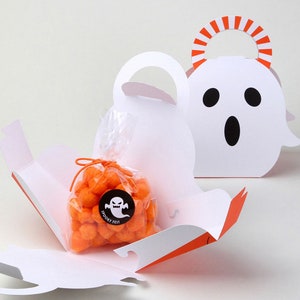 May include: Two white ghost-shaped paper treat boxes with orange and white striped handles. One box is open, revealing a clear plastic bag filled with orange spherical snacks, tied with an orange string and a black sticker labeled Spooky Fest.