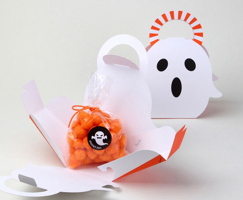 May include: Two white ghost-shaped paper treat boxes with orange and white striped handles. One box is open, revealing a clear plastic bag filled with orange spherical snacks, tied with an orange string and a black sticker labeled Spooky Fest.