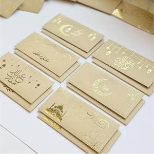 May include: A collection of beige paper envelopes with gold foil designs and Arabic calligraphy. The envelopes feature crescent moons, mosque illustrations, and floral patterns. The envelopes are stacked in neat piles, suggesting a festive or celebratory theme.