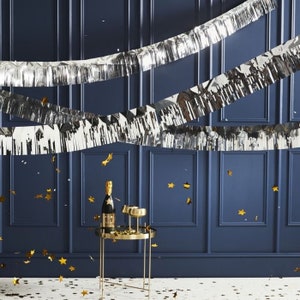 May include: Three silver tinsel garlands hang against a dark blue wall. Gold confetti is scattered on the floor in front of a gold metal side table with two champagne glasses and a bottle of champagne.