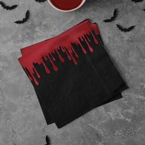 May include: Black and red paper napkins with a dripping blood design. The napkins are folded in half and are on a gray surface.