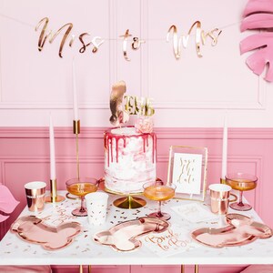 May include: A pink and white cake with gold glitter and a 'Bride to Be' topper. The cake is on a white cake stand and is surrounded by rose gold party supplies, including plates, cups, and a banner that says 'Miss to Mrs'.