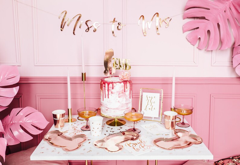 May include: A pink and white cake with gold glitter and a 'Bride to Be' topper. The cake is on a white cake stand and is surrounded by rose gold party supplies, including plates, cups, and a banner that says 'Miss to Mrs'.