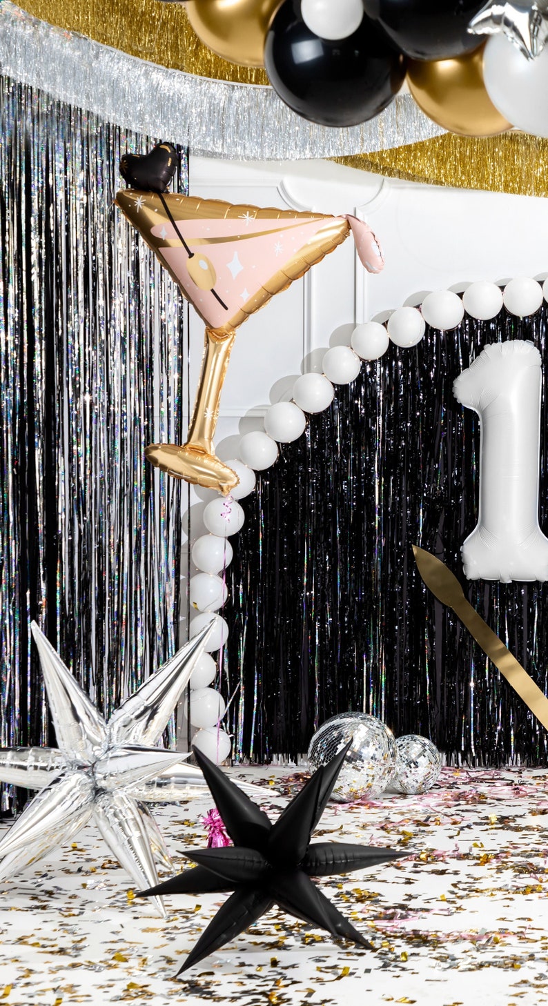 May include: A black and silver party backdrop with a silver star, a black star, and a gold and pink martini glass balloon. The backdrop is decorated with silver and gold fringe and white balloons. There is confetti on the floor.