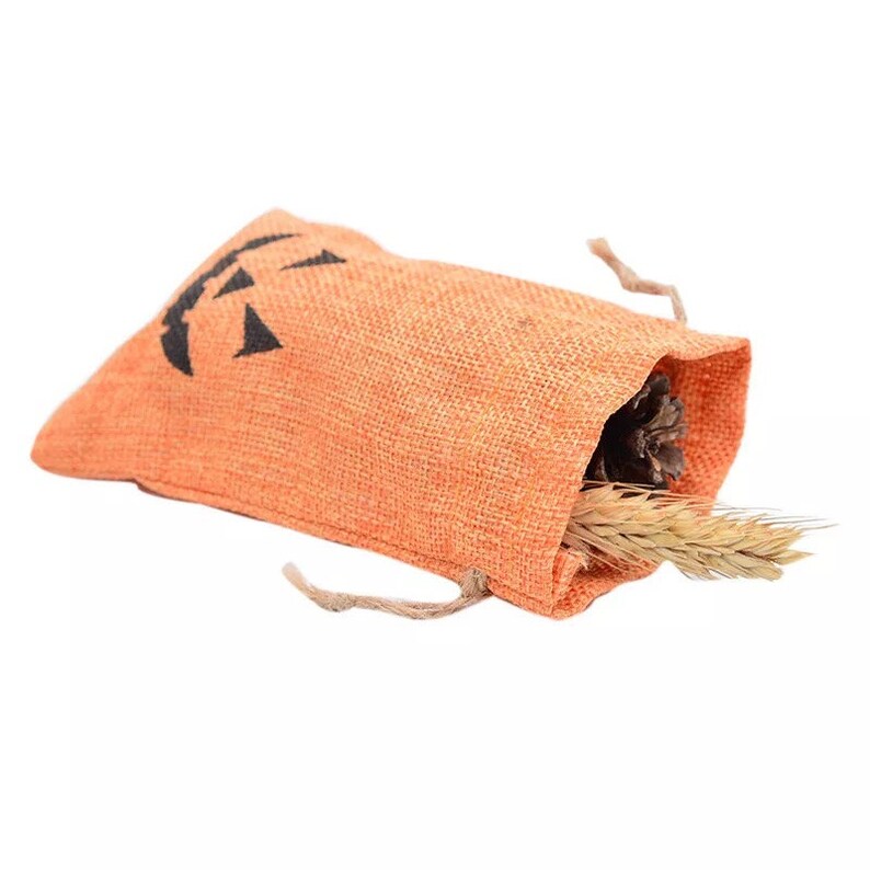 May include: An orange burlap drawstring pouch featuring a black jack-o'-lantern face print. The bag is partially open, revealing a dark brown pinecone and a stalk of dried wheat protruding from the top opening of the textured fabric sack.