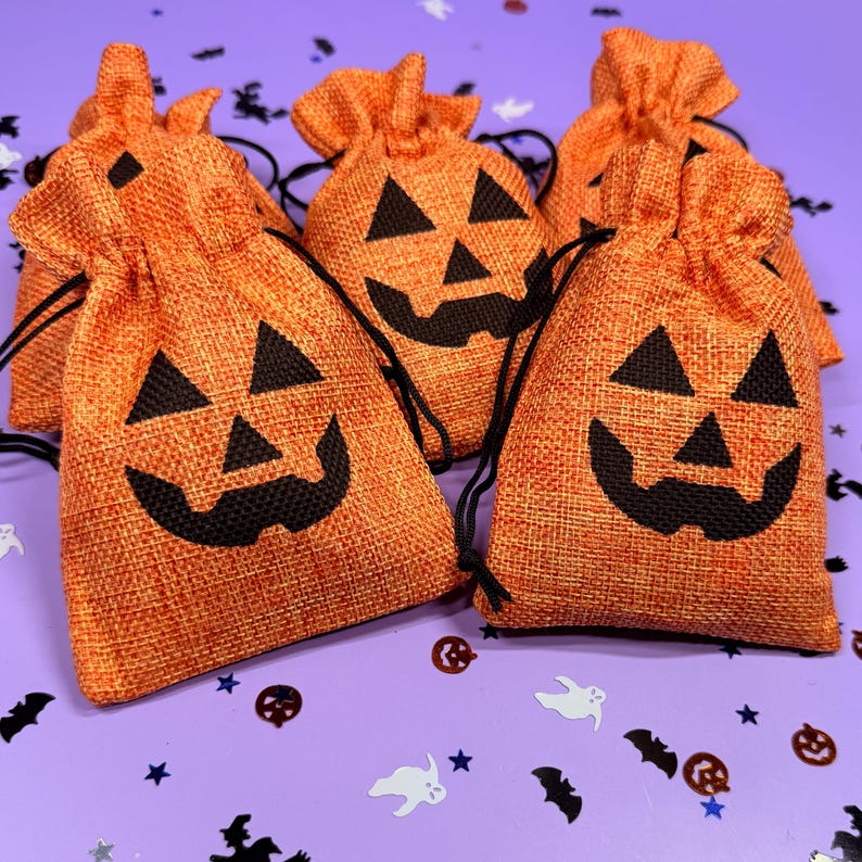 May include: A collection of orange burlap drawstring bags featuring a black jack-o'-lantern face print. Each small pouch has a textured fabric surface and black cord ties at the top, arranged together to display the festive pumpkin designs.