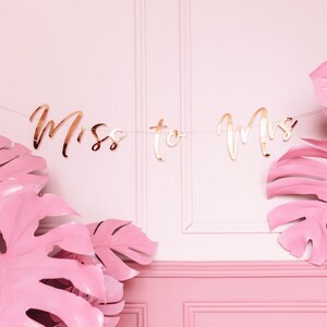 May include: A pink and gold banner with the text "Miss to Mrs" hanging between two large pink monstera leaves.