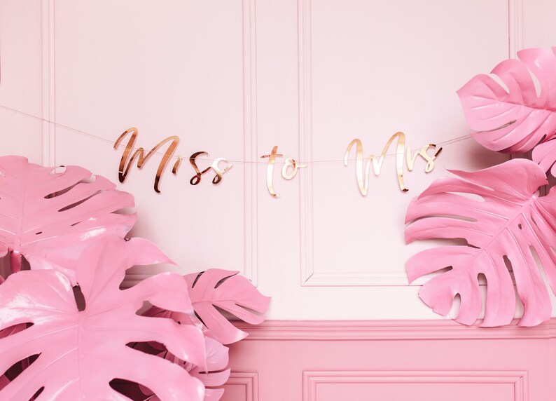 May include: A pink and gold banner with the text "Miss to Mrs" hanging between two large pink monstera leaves.