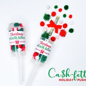May include: Two clear plastic push-pop containers filled with red, green, and white pom-poms. One container has a rolled-up bill with a red bow. The containers have labels that read "Christmas Cash-fetti Holiday Push-Pop."