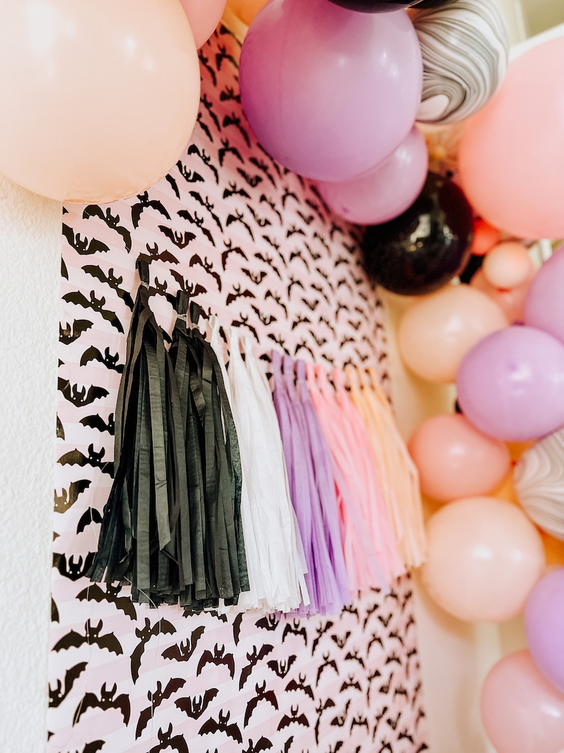 Op de afbeelding: Een roze en zwarte achtergrond met vleermuis-thema met zwarte, witte, paarse en roze tissue-kwastjes die aan de onderkant hangen. De achtergrond is versierd met een herhalend patroon van zwarte vleermuizen op een roze achtergrond. De achtergrond is omringd door een verscheidenheid aan ballonnen in roze, paars, zwart en wit.