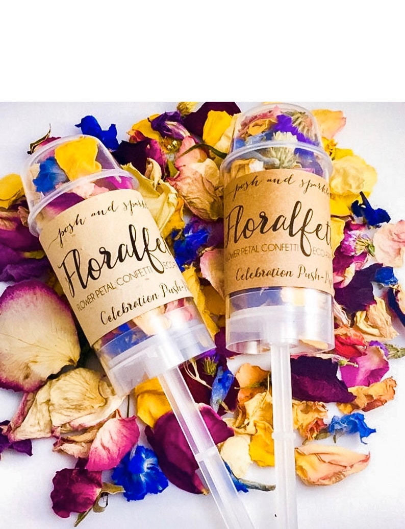 K&ouml;nnte beinhalten: Zwei transparente Konfettikanonen aus Kunststoff, gef&uuml;llt mit getrockneten Bl&uuml;tenbl&auml;ttern. Die Kanonen haben braune Papieretiketten mit der Aufschrift "posh and sparkle Floralfet Flower Petal Confetti Eco Celebration Push-Pop".