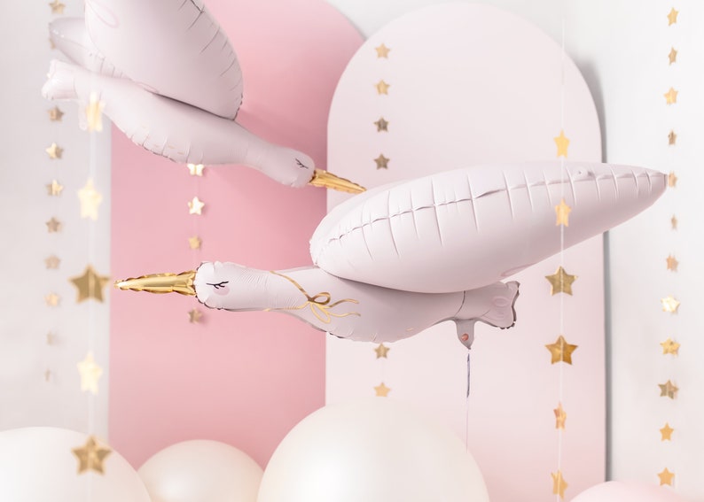 May include: Light pink stork-shaped foil balloons with gold beaks and accents float in a room decorated with gold star garlands and white and pink balloons. The background features pink arches.