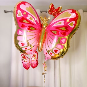 May include: A large pink and gold butterfly balloon with a smaller pink butterfly balloon attached to the bottom. The balloons are decorated with hearts and gold accents.