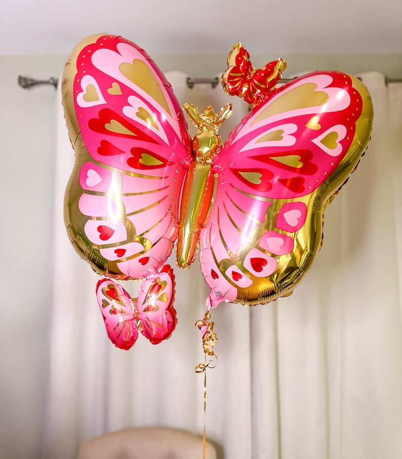 May include: A large pink and gold butterfly balloon with a smaller pink butterfly balloon attached to the bottom. The balloons are decorated with hearts and gold accents.