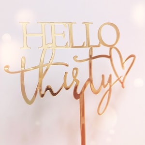 以下が含まれることがあります： 「Hello Thirty」とハートが書かれたゴールドのグリッターケーキトッパー