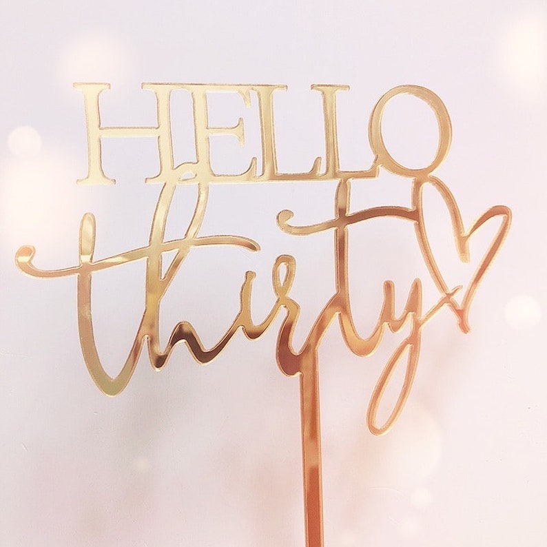 以下が含まれることがあります： 「Hello Thirty」とハートが書かれたゴールドのグリッターケーキトッパー