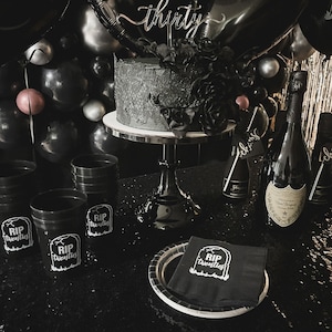 May include: A black-themed birthday celebration featuring a cake, balloons, and champagne bottles. The cake is adorned with black floral decorations and a 'thirty' cake topper. Black cups and napkins display the text 'RIP Twenties'.