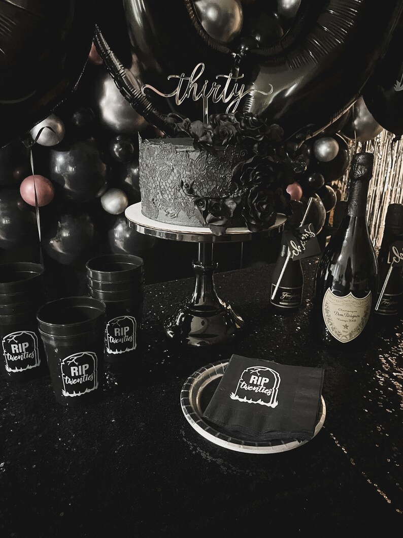 May include: A black-themed birthday celebration featuring a cake, balloons, and champagne bottles. The cake is adorned with black floral decorations and a 'thirty' cake topper. Black cups and napkins display the text 'RIP Twenties'.