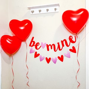 May include: Red heart-shaped balloons and a red banner with the text "be mine" hanging on a white wall. The banner is decorated with pink glitter hearts.