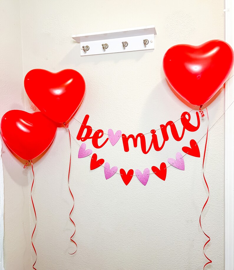 May include: Red heart-shaped balloons and a red banner with the text "be mine" hanging on a white wall. The banner is decorated with pink glitter hearts.