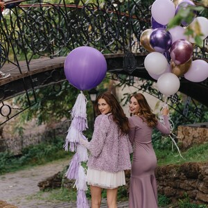 May include: Two women in purple dresses stand under a bridge decorated with purple, gold, and white balloons. The woman on the left is holding a large purple balloon with a white and purple tassel.