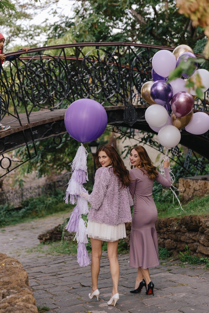 May include: Two women in purple dresses stand under a bridge decorated with purple, gold, and white balloons. The woman on the left is holding a large purple balloon with a white and purple tassel.