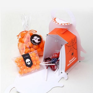 May include: Two clear plastic bags filled with orange spherical candies, one featuring a ghost sticker labeled Spooky Fest and the other a skull sticker. An orange cardboard box with a spiderweb and spider print labeled Happy Halloween.