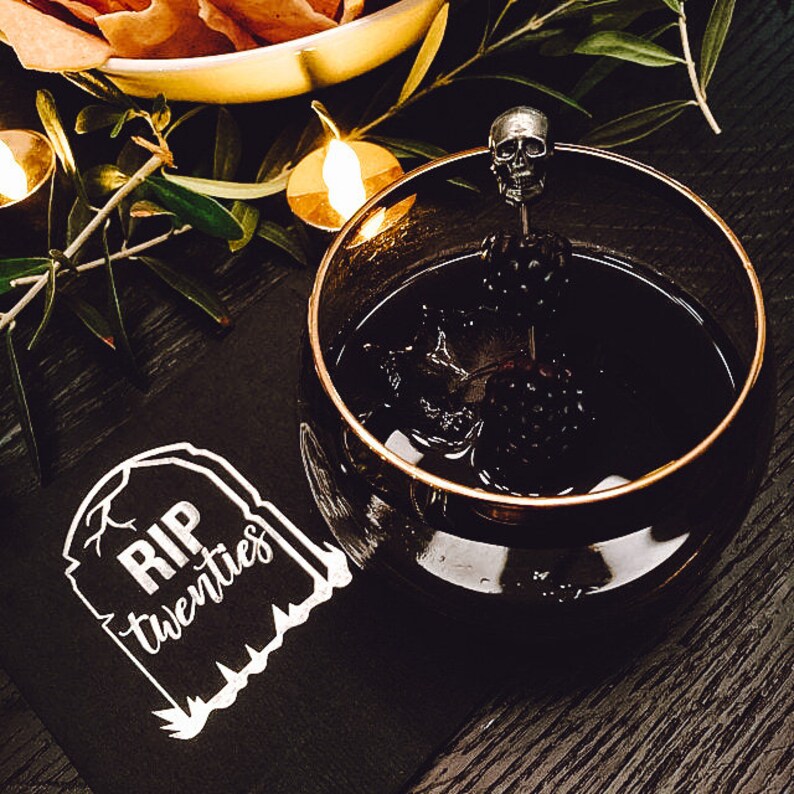 May include: A black cocktail in a dark glass with a gold rim, garnished with blackberries and a skull-topped stirrer. A black napkin with a tombstone design and the text "RIP Twenties" is in the foreground. The scene has a Halloween theme.